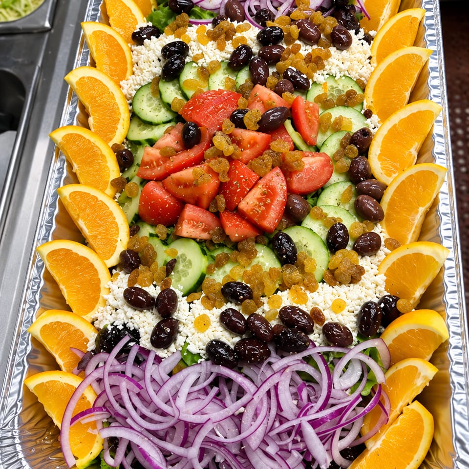 Large Mediterranean salad platter with tomatoes, cucumbers, feta, olives, raisins, red onions, greens, and orange wedges.