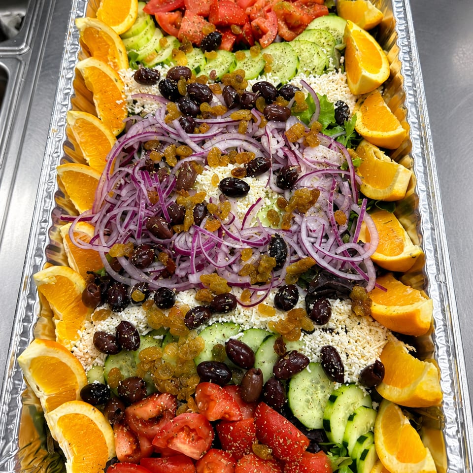 Large foil tray filled with layered salad, tomatoes, cucumbers, red onions, olives, raisins, feta, and orange wedges.