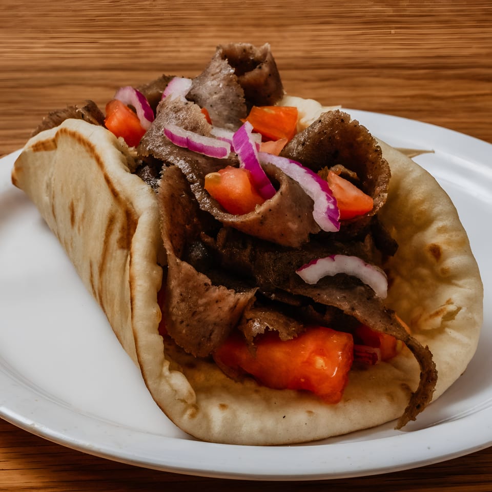 A gyro with meat, tomatoes, and red onions on a white plate, on a wooden table.