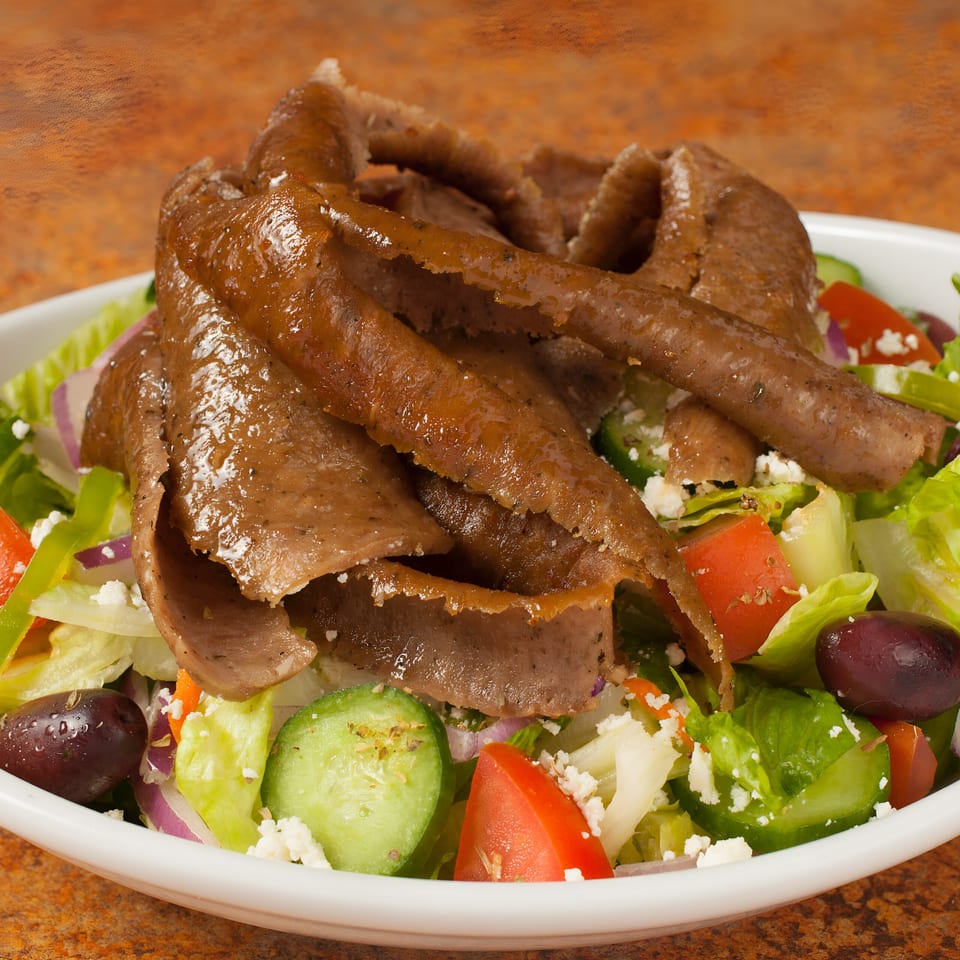 A white bowl filled with a fresh salad topped with gyro meat, feta, olives, and vegetables.