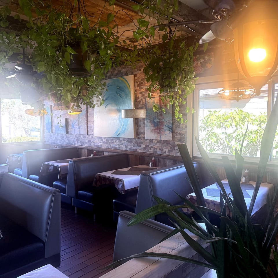 Restaurant interior with booths, blue wave artwork, and trailing plants near the windows.