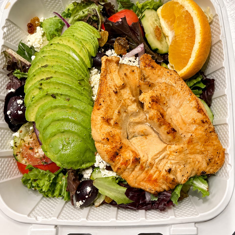 Grilled chicken salad with avocado, mixed greens, cucumber, tomato, feta cheese, raisins, olives, and an orange wedge in a takeout container.