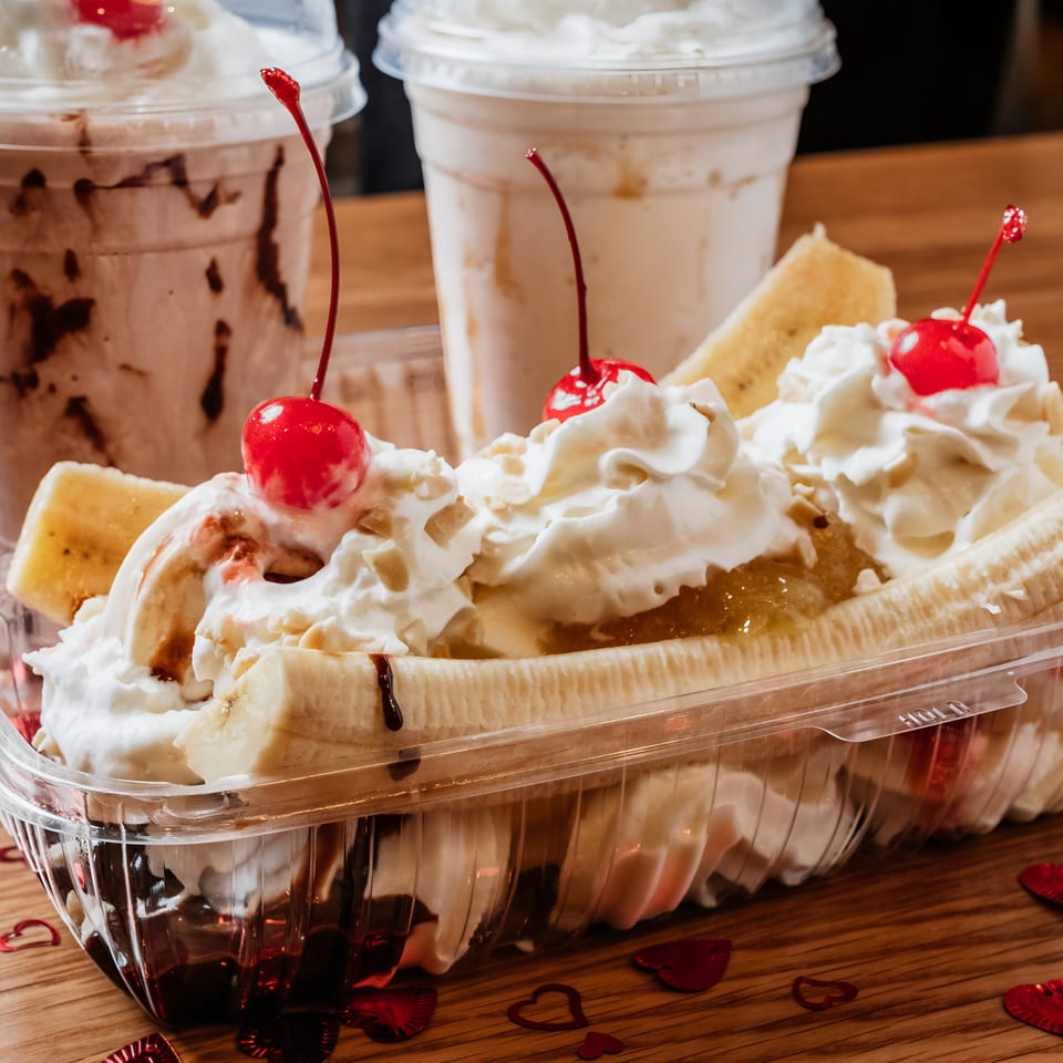 Banana split with whipped cream, cherries, and two milkshakes on a wooden table with heart confetti.